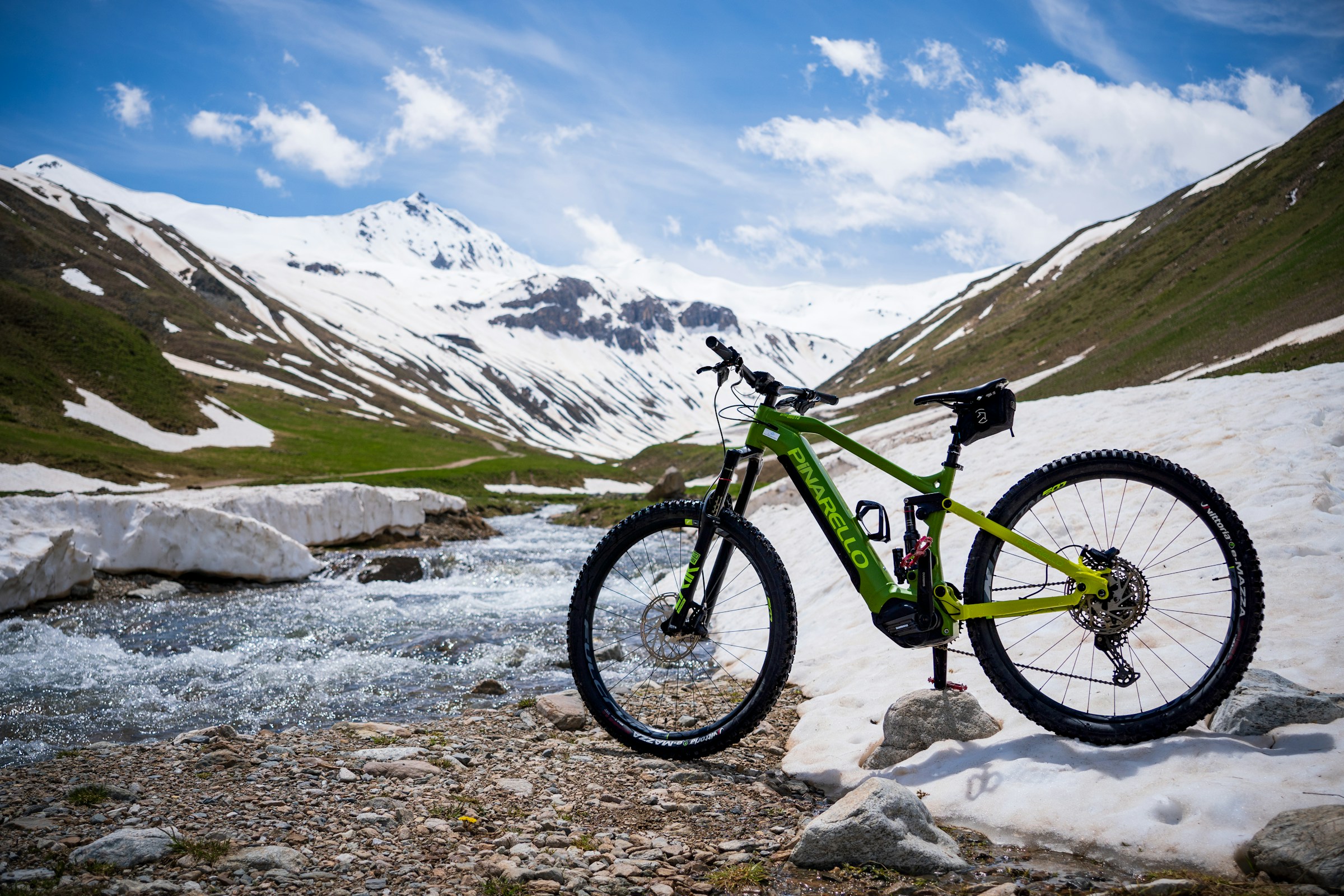 Mountain bike electric (E-MTB) marca Pinarello, verde, staționat pe o zonă cu zăpadă și pietriș lângă un pârâu de munte, cu vârfuri înzăpezite în fundal.